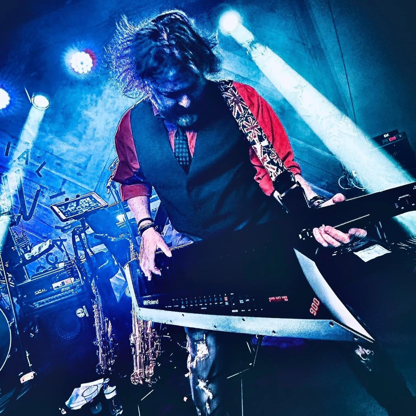 Andrew Doss on a blue-lit stage leaning into a black Roland keytar slung from a Union Jack strap, wearing a red shirt and dark vest and tie, with a saxophone on a stand beside him